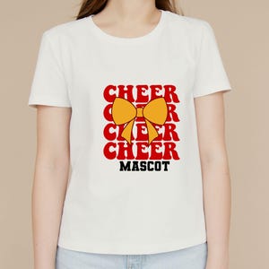 May include: White t-shirt with the word "CHEER" in red, repeated three times, and a yellow bow graphic. The word "MASCOT" is printed in black below the word "CHEER".