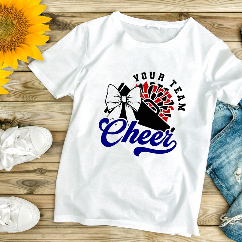 May include: White t-shirt with a cheerleading design. The graphic includes a bow, pom-poms, and the words "Your Team Cheer" in black and blue. The shirt is displayed on a wooden surface with a sunflower and white sneakers.
