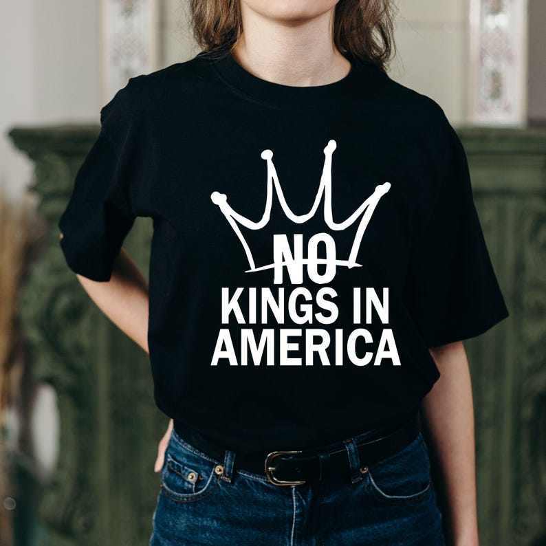 K&ouml;nnte beinhalten: Schwarzes T-Shirt mit wei&szlig;em Kronen-Design und dem Text "NO KINGS IN AMERICA". Das Shirt hat kurze &Auml;rmel und ist aus weichem Stoff. Der Aufdruck ist mittig auf der Vorderseite platziert.