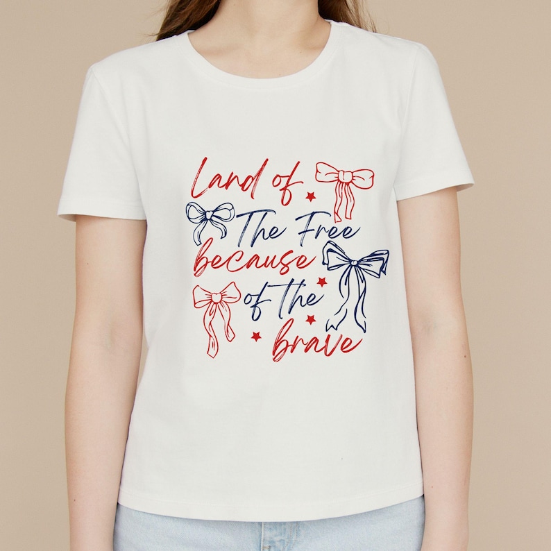 May include: White t-shirt with the text "Land of the Free because of the brave" in red and blue script. The design includes bow illustrations and stars. A casual, patriotic tee.