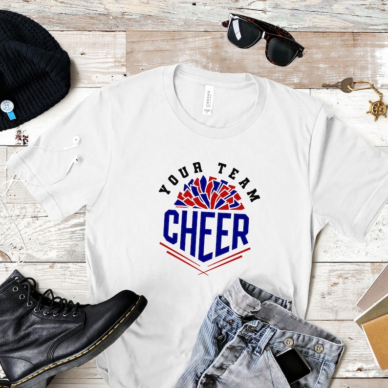 May include: White t-shirt with the text "YOUR TEAM CHEER" in red and blue. The shirt is displayed with black boots, sunglasses, and a black beanie on a wooden surface.