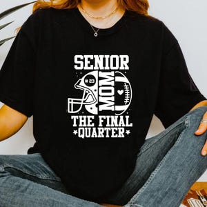 以下が含まれることがあります： 白のグラフィックデザインが施された黒いTシャツ。デザインには、フットボールのヘルメット、フットボール、そして「SENIOR MOM THE FINAL QUARTER」という言葉が含まれています。「23」の数字がヘルメットの中にあります。