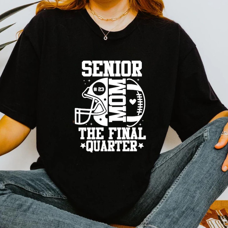 以下が含まれることがあります： 白のグラフィックデザインが施された黒いTシャツ。デザインには、フットボールのヘルメット、フットボール、そして「SENIOR MOM THE FINAL QUARTER」という言葉が含まれています。「23」の数字がヘルメットの中にあります。