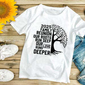 May include: White t-shirt with a black graphic of a tree and the text "2025 Family Reunion Our Roots Run Deep Our Love Runs Deeper." The shirt is laid flat on a wooden surface with a sunflower and white sneakers.