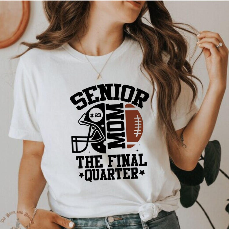 以下が含まれることがあります： 白のTシャツ。黒と茶色のフットボールをテーマにしたグラフィック。グラフィックには、フットボールのヘルメット、フットボール、そして「SENIOR MOM #23 THE FINAL QUARTER」という文字が含まれています。Tシャツはクルーネックです。