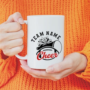 May include: White ceramic mug with a black and red cheerleading design. The design includes a megaphone, pom-poms, and a football, with the word "Cheer" in red script. The mug also has the words "Team Name" in a black, bold font.