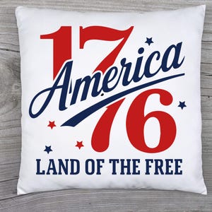 May include: White square pillow with the text "1776 America" in red and blue, with star accents and the words "LAND OF THE FREE" below. The pillow is on a wooden surface.