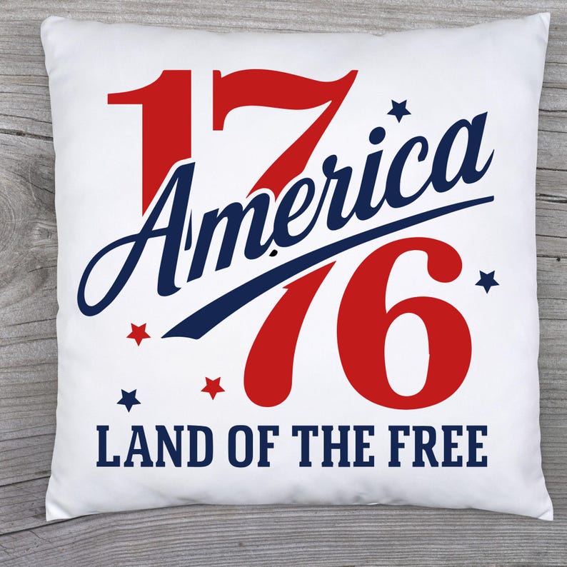 May include: White square pillow with the text "1776 America" in red and blue, with star accents and the words "LAND OF THE FREE" below. The pillow is on a wooden surface.