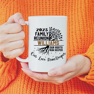May include: White ceramic mug with black text that reads "2025 Family Reunion Williams Our Roots Run Deeper Our Love Runs Deeper" and a tree graphic. The mug is held by a person wearing an orange sweater.