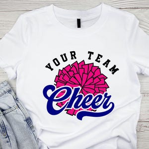 May include: White t-shirt with the words "YOUR TEAM Cheer" in black and blue. A pink pom-pom graphic is centered on the shirt. The shirt is paired with blue jeans.