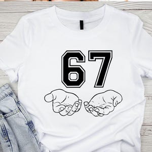 K&ouml;nnte beinhalten: Wei&szlig;es T-Shirt mit der Zahl "67" in schwarzer, college-artiger Schrift. Unter den Zahlen befinden sich zwei offene H&auml;nde, ebenfalls in Schwarz. Das Design ist mittig auf dem Shirt platziert.