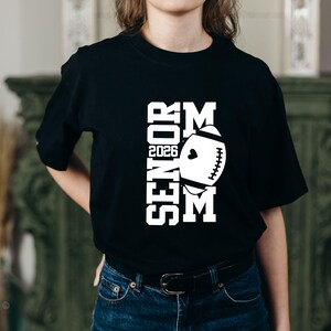 以下が含まれることがあります： 白文字で「SENIOR MOM 2026」と白いアメフトのグラフィックがプリントされた黒いTシャツ。クルーネックのTシャツで、青いジーンズと合わせて着用されています。デザインは前面の中央に配置されています。
