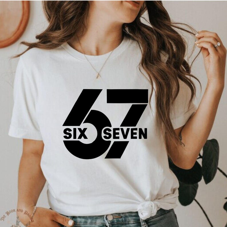 K&ouml;nnte beinhalten: Wei&szlig;es T-Shirt mit einem gro&szlig;en schwarzen Aufdruck der Zahlen "67" und den Worten "SIX SEVEN" in fetter Schrift. Das T-Shirt hat einen Rundhalsausschnitt und ein schlichtes Design.