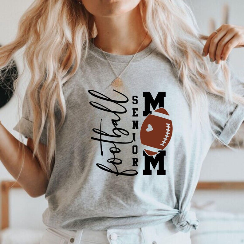 以下が含まれることがあります： 「Football Senior Mom」の文字が黒い筆記体とブロック体でプリントされた、ライトグレーのTシャツ。茶色のフットボールと白いハートが「MOM」の文字の中央に配置されています。Tシャツは半袖で、クルーネックです。