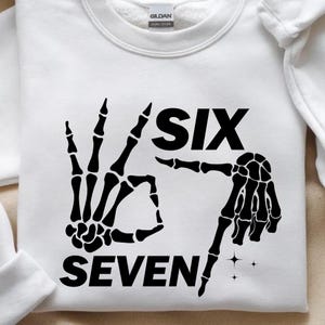 K&ouml;nnte beinhalten: Wei&szlig;es Sweatshirt mit einer schwarzen Skeletthand-Grafik, die die Zahlen sechs und sieben bildet. Das Wort "SIX" steht &uuml;ber der Skeletthand, und "SEVEN" steht darunter. Das Sweatshirt hat einen Rundhalsausschnitt.