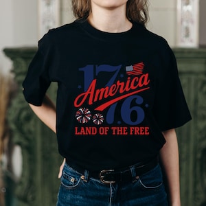 May include: Black t-shirt with a patriotic design. The design includes the text "1776 America" in red and blue, with an American flag and fireworks graphic. The phrase "Land of the Free" is also included.
