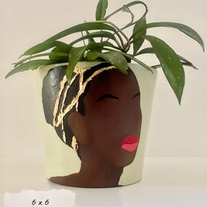 May include: A decorative ceramic planter featuring a painted portrait of a woman with dark skin, black hair, and gold accents. The woman has red lips and closed eyes. The planter is filled with green leafy plants. The planter measures 6 x 6 inches.