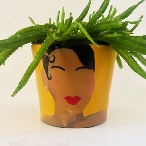 May include: A yellow painted ceramic planter with a stylized face design, holding a green plant. The planter is approximately 6 x 6 inches. The face has black hair, red lips, and a gold earring. The plant has long, thin, green stems.