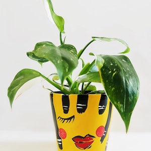 May include: A yellow painted ceramic plant pot with a face design. The pot features black stripes, closed eyes, red blush, and red lips. The pot holds a green plant with large, speckled leaves.