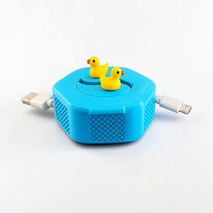May include: A bright blue hexagonal cable organizer featuring two small yellow rubber ducks on the top surface. The device has a textured grid pattern on its sides and includes two white USB charging cables extending from opposite ends of the unit.