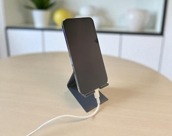 Puede incluir: Un smartphone gris oscuro se apoya en un soporte a juego, conectado a un cable de carga blanco. El soporte está inclinado, proporcionando una vista clara de la pantalla del teléfono. El conjunto está sobre una mesa de madera clara, sugiriendo una estética moderna.