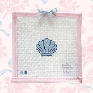 Puede incluir: Un bordado cuadrado con un diseño de concha de color azul claro sobre un fondo blanco. El bordado está enmarcado en rosa y adornado con un lazo azul claro. El fondo es un estampado floral rosa.