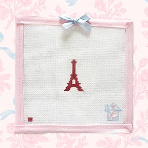 May include: A square, cross-stitched artwork featuring a red Eiffel Tower on a white background. The piece is framed in pink with a light blue bow at the top. A small house and square are also cross-stitched.