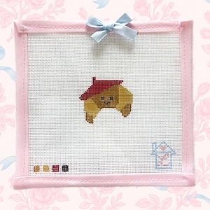 May include: A cross-stitch piece featuring a cartoon croissant with a red roof, set within a pink frame. A light blue bow adorns the top. The background is a floral pattern with a light pink color.