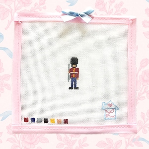 May include: Cross-stitch art featuring a British Royal Guard in a red jacket and black hat, holding a rifle. The artwork is framed in pink with a light blue bow at the top. A small house and color swatches are at the bottom.