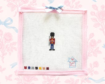 May include: Cross-stitch art featuring a British Royal Guard in a red jacket and black hat, holding a rifle. The artwork is framed in pink with a light blue bow at the top. A small house and color swatches are at the bottom.