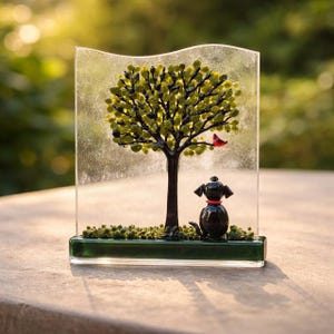 May include: A glass art piece featuring a tree with green leaves and a red bird. A black dog with a red collar sits below the tree. The art piece is set on a green base and encased in clear glass.