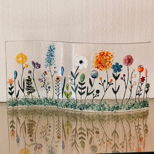 May include: A glass art piece depicting a field of flowers with various colors, including yellow, orange, white, pink, blue, and green. The flowers are made of glass and are arranged in a wavy pattern. The piece is set against a white background.
