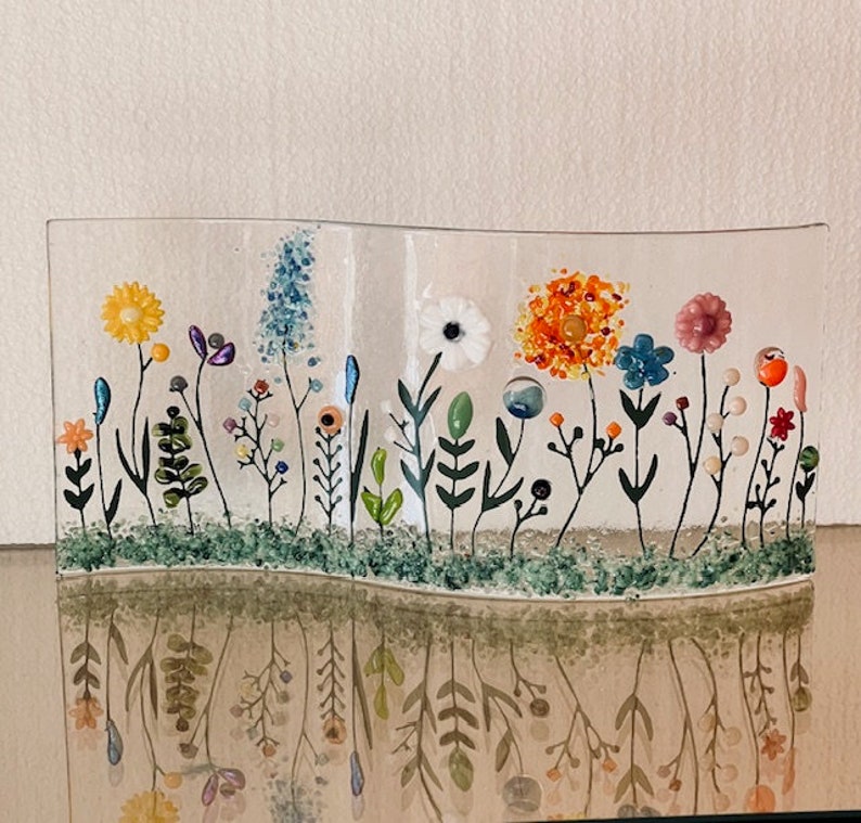 May include: A glass art piece depicting a field of flowers with various colors, including yellow, orange, white, pink, blue, and green. The flowers are made of glass and are arranged in a wavy pattern. The piece is set against a white background.