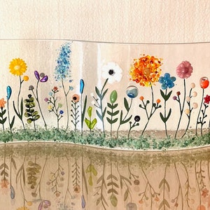May include: A glass art piece depicting a field of flowers in various colors, including yellow, orange, pink, blue, and green. The flowers are arranged in a curved line, creating a sense of movement. The piece is set against a white background.