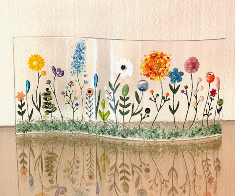 May include: A glass art piece depicting a field of flowers in various colors, including yellow, orange, pink, blue, and green. The flowers are arranged in a curved line, creating a sense of movement. The piece is set against a white background.
