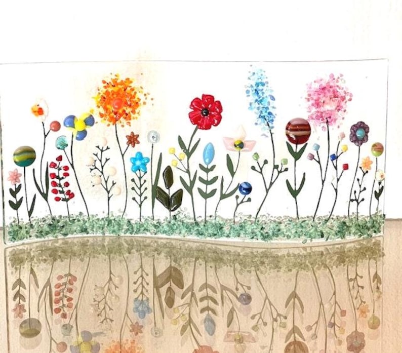 May include: A glass art piece depicting a colorful garden with flowers, leaves, and stems. The flowers are made of glass beads and the stems are made of black wire. The garden is set against a white background and is reflected in a mirror-like surface below.