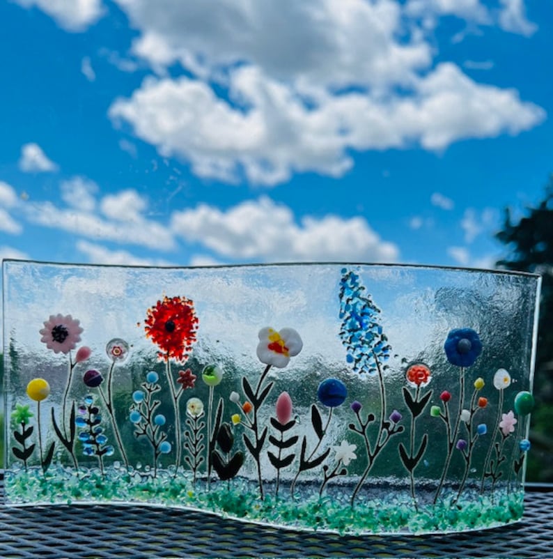 May include: A glass art piece depicting a field of colorful flowers with black stems and green grass. The flowers are made of glass and have a variety of colors, including red, blue, yellow, pink, and white.