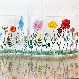 May include: A glass art piece depicting a field of colorful flowers with green stems and leaves. The flowers are made of glass and are arranged in a row across the piece. The piece is set on a white background.