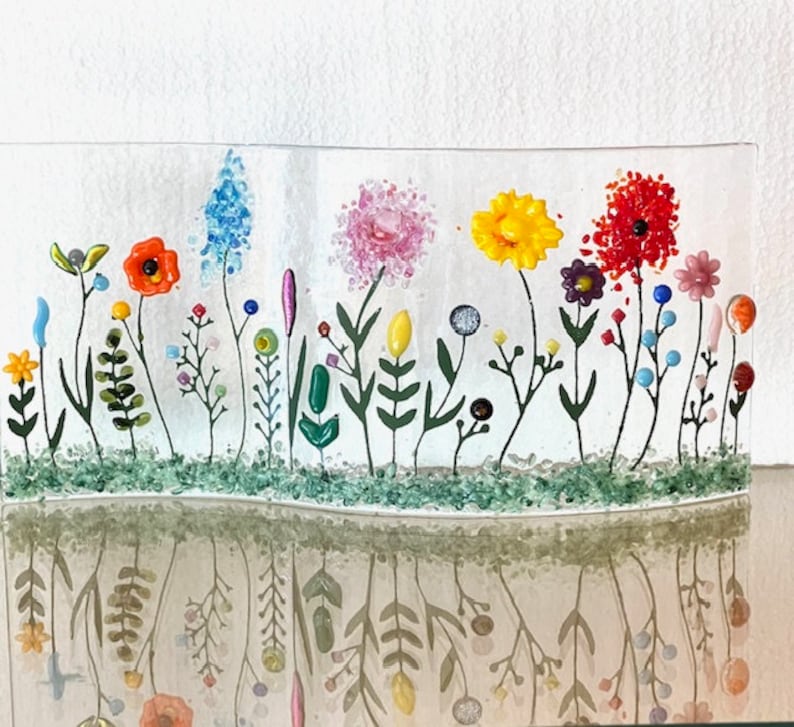 May include: A glass art piece depicting a field of colorful flowers with green stems and leaves. The flowers are made of glass and are arranged in a row across the piece. The piece is set on a white background.