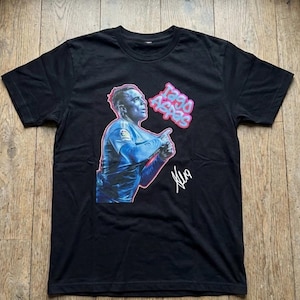 May include: Black t-shirt featuring a graphic of a soccer player in a blue jersey with the text "Iago Aspas" in pink and white bubble letters. The shirt also includes a signature in white.