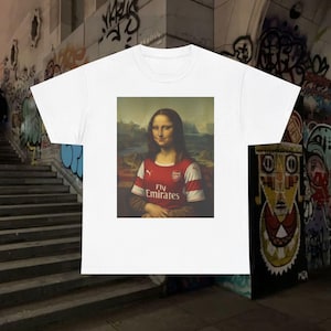 Puede incluir: Camiseta blanca con una parodia de la Mona Lisa con una camiseta de fútbol roja y blanca con el texto "Fly Emirates". El fondo presenta arte de graffiti.