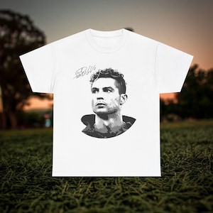 May include: White t-shirt featuring a black and white portrait of a soccer player with a signature above the image. The portrait is a detailed illustration of the player's face and upper body. The shirt is made of a soft material.