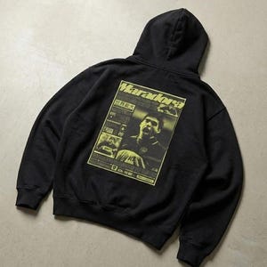 Può includere: Felpa con cappuccio nera con grafica gialla sul retro. La grafica è un design in stile giornale con il nome "Maradona" e immagini di un calciatore. La felpa ha un cappuccio e maniche lunghe.