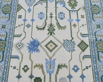 May include: A rectangular area rug featuring a cream-colored background with a repeating geometric and floral pattern in shades of light blue, dark blue, and olive green. The design includes symmetrical motifs and a decorative border.