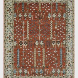 Puede incluir: Una alfombra rectangular con un campo de color rojo terracota profundo y un patrón repetitivo de motivos florales y arbóreos estilizados en tonos azul, crema y verde. La alfombra está enmarcada por un borde ancho de color crema.