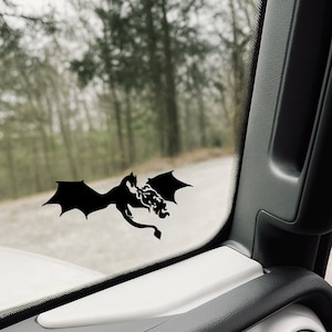 May include: A black vinyl decal of a dragon with outstretched wings is affixed to a vehicle window. The dragon is silhouetted against a blurred outdoor background of trees and a road. The decal is on the inside of the window.