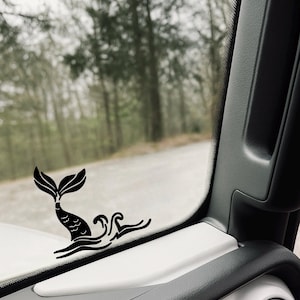 May include: A black mermaid tail and wave decal is affixed to the interior of a vehicle. The decal is positioned on the white dashboard near the window, with a blurred forest and road visible in the background.
