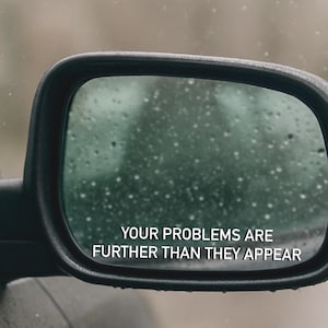 May include: A black side-view mirror with the text "YOUR PROBLEMS ARE FURTHER THAN THEY APPEAR" in white. The mirror's glass is covered in water droplets, suggesting rain. The background is blurred, with a muted color palette.