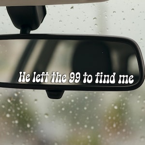 Può includere: Specchietto retrovisore nero di un'auto con la scritta bianca "He left the 99 to find me" in un font retrò. Lo sfondo sfocato mostra una giornata piovosa attraverso il parabrezza.