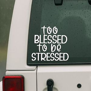 Op de afbeelding: Witte vinyl sticker met de tekst "too BLESSED to be STRESSED" in een handgeschreven lettertype. De sticker zit op de achterruit van een wit voertuig, met een rood achterlicht zichtbaar.